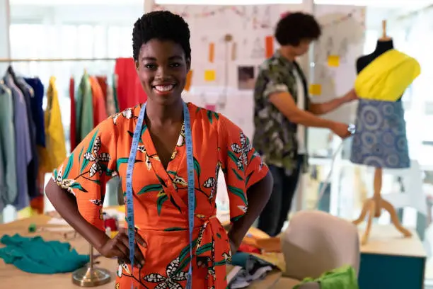 Portrait of young pretty African american female fashion designer standing with hands on hip in design studio. Mixed race man working in the background. This is a casual creative start-up business office for a diverse team