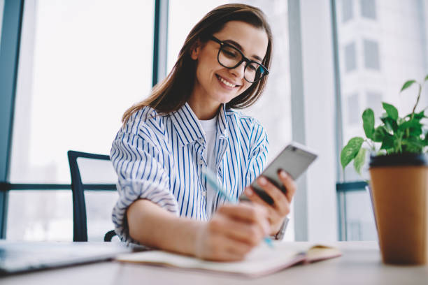 istock Young millennial girl in eyewear reading income message on smartphone while writing homework, smiling female employee checking email making notes of creative ideas for article publication in blog 1161359556