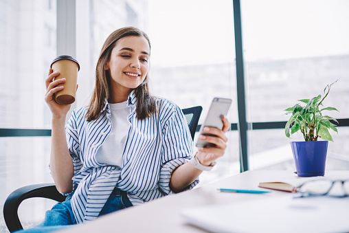 Cheerful young woman watching video in social networks on smartphone during break holding coffee cup, smiling  hipster girl sending text message via mobile phone sitting at desktop in office