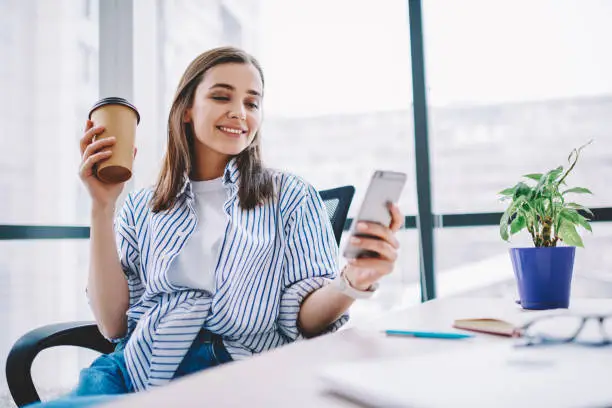 Photo of Cheerful young woman watching video in social networks on smartphone during break holding coffee cup, smiling  hipster girl sending text message via mobile phone sitting at desktop in office