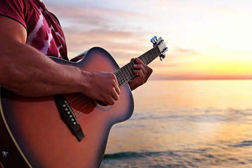 Young man wearing purple tie dye t-shirt playing dreadnought parlor acoustic guitar on beach at beautiful sunset time. Fit guitarist w/ sunburst instrument by the sea. Background, copy space, close up