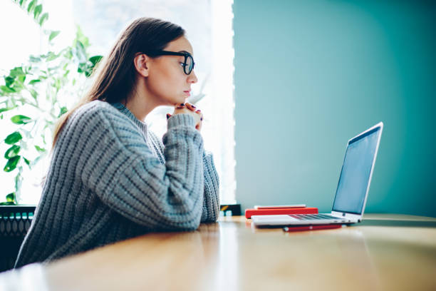 istock Pensive young woman in optical eyeglasses watching interesting webinar of learning foreign languages during remote education in coworking.Concentrated hipster girl studying at laptop computer in college 1161370412