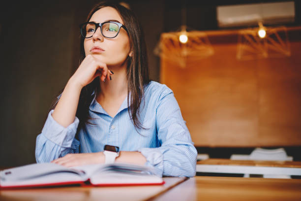 Pensive young woman in eyewear pondering solving problem with business project in cafe interior, female student preparing for examination analyzing information from notebook spending time in campus mujer joven pensativa en gafas que meditan resolviendo el problema con el proyecto de negocios en el interior del café, estudiante femenina que se prepara para el examen analizando la información de la libreta pasando tiempo en el campus