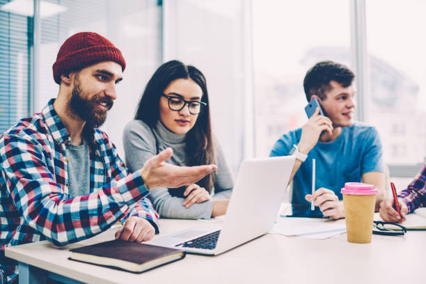 istock Group of skilled it professionals creating website togetherness using computer, young hipster guy updating software for online work while female colleague sitting next to him at university campus 1161376003