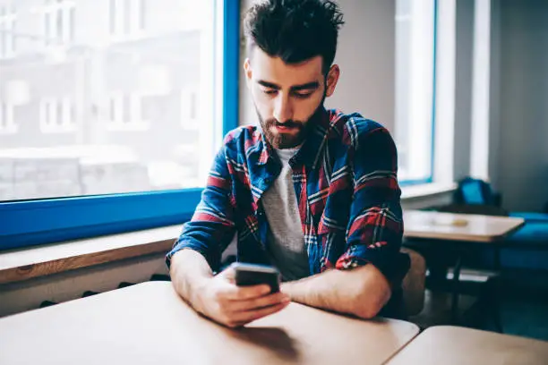 Bearded hipster guy dressed in casual wear messaging in online chat on modern smartphone using free 4G internet connection sitting at desktop in college.Student updating profile in social networks