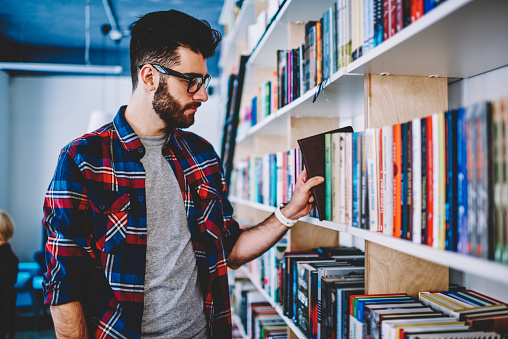 Pensive bearded student in eyeglasses standing near big bookshelf and choosing literature for coursework.Smart young man in eyewear holding book in hand preparing for exam in educational library