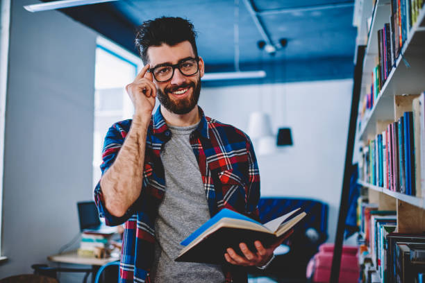 istock Smiling young man in eyewear holding interesting book in university campus and looking at camera, happy successful male student in trendy apparel spending free time on autodidact enjoying  literature 1161383436