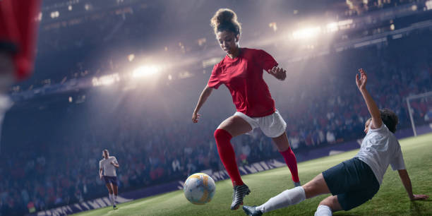 Professional Woman Soccer Player Jumping Over Sliding Tackle During Match Mid action image of a professional woman soccer player in mid air, jumping over an opposition player's sliding tackle. The footballer is playing on a grass soccer pitch in a generic floodlit stadium full of spectators.  Both players wear generic red and white, and dark blue and white soccer uniform. womens soccer stock pictures, royalty-free photos & images