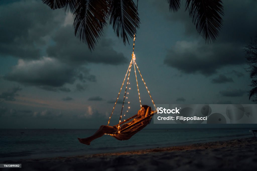 Young adult woman relaxing on a swing in a tropical paradise Night Stock Photo