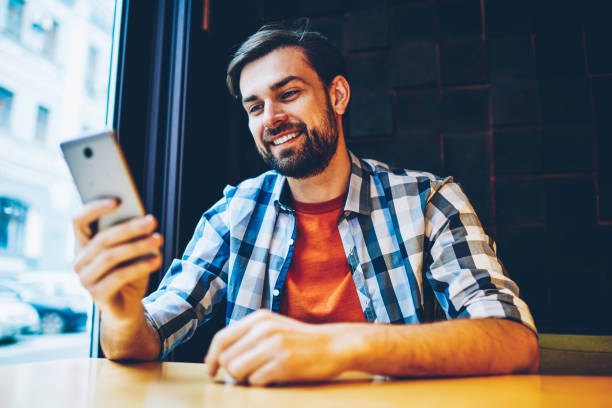 istock Successful bearded young man messaging in online chat on modern smartphone via 4G internet sitting in coworking space.Positive hipster blogger reading notification with good news on cellular device 1161405316