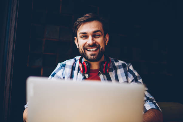 istock Portrait of at successful bearded male freelancer smiling at camera while working remotely at laptop device.Cheerful it developer with headphones sitting in coworking space at netbook with 4G internet 1161405358