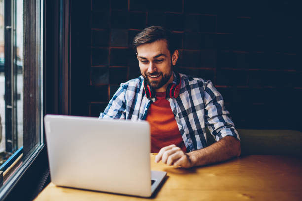 istock Cheerful bearded student with headphones on neck watching funny tutorial on website on modern netbook device connected to wireless internet.Positive young man freelancer reading news in blog on laptop 1161405361