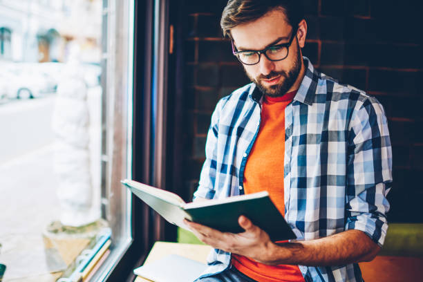 istock Concentrated smart student in eyeglasses for vision correction reading literature book preparing for upcoming exams in university.Pensive intelligent young man enjoying bestseller in free time 1161405395