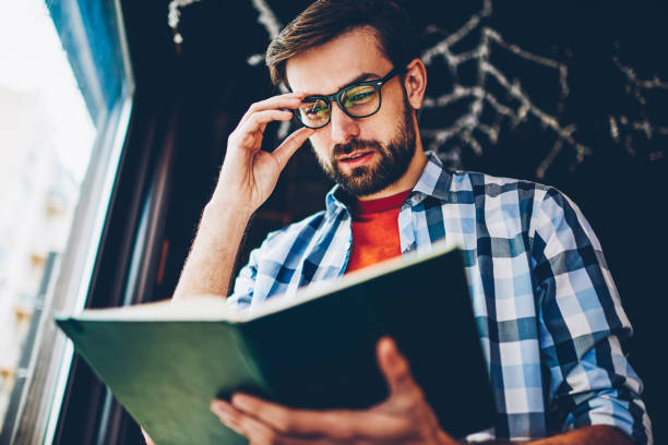 istock Concentrated smart young man with beard and optical eyeglasses searching useful information in textbook for coursework.Pensive hipster guy reading book preparing for studying seminar in coworking 1161405406