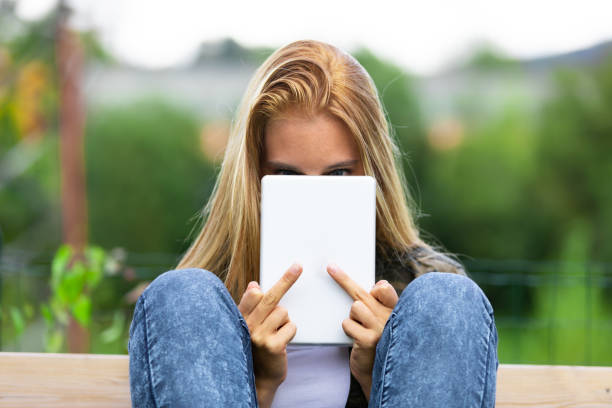 Young woman making a rude sign with her finger Young woman making a rude derogatory sign with her middle fingers as she hides behind a handheld tablet computer while relaxing outdoors in the garden on a bench derogatory stock pictures, royalty-free photos & images