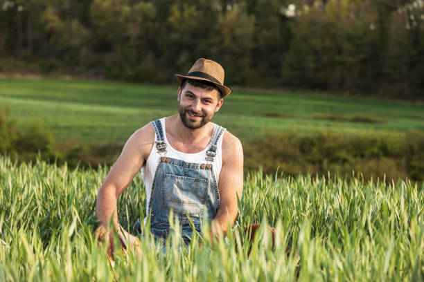istock Bearded smiling young man wearing a hat sitting on field while looking camera 1161409694