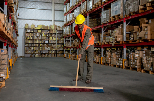 Black young man working at a distriution warehouse sweeping the floor - Business industry concepts