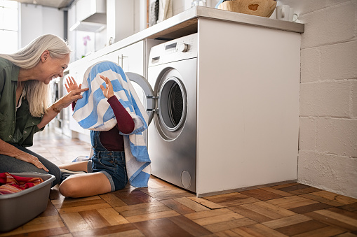 Child hiding in blue and striped towel scaring mature woman near washing machine at home. Smiling old grandmother and funnny little girl playing with fresh laundry clothes on floor. Grandchild plays the ghost with her head covered by a towel.