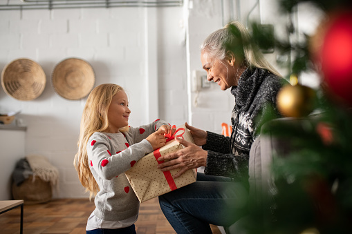 Happy senior woman giving christmas surprise to little girl at home. Excited granddaughter unpacking present under christams tree with old granny in sweater. Grandmother giving xmas gift to grandchild.