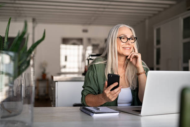 Senior woman using laptop and smartphone Happy senior woman holding smartphone and laptop daydreaming while looking away. Successful stylish old woman working at home while thinking about a good future. Cheerful fashionable lady entrepreneur wearing cool eyeglasses. mid adult stock pictures, royalty-free photos & images