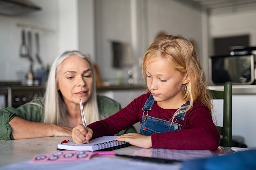 Beautiful girl child with senior woman writing on notebook. Cute little schoolgirl writing in notebook. Granddaughter with grandmother studying at home while sitting on table and doing homework together.