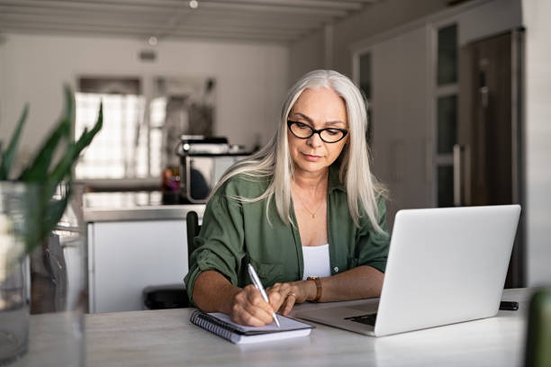 Senior fashionable woman working at home Senior stylish woman taking notes in notebook while using laptop at home. Old freelancer writing details on book while working on laptop in living room. Focused cool lady writing notary in notepad. white hair photos stock pictures, royalty-free photos & images