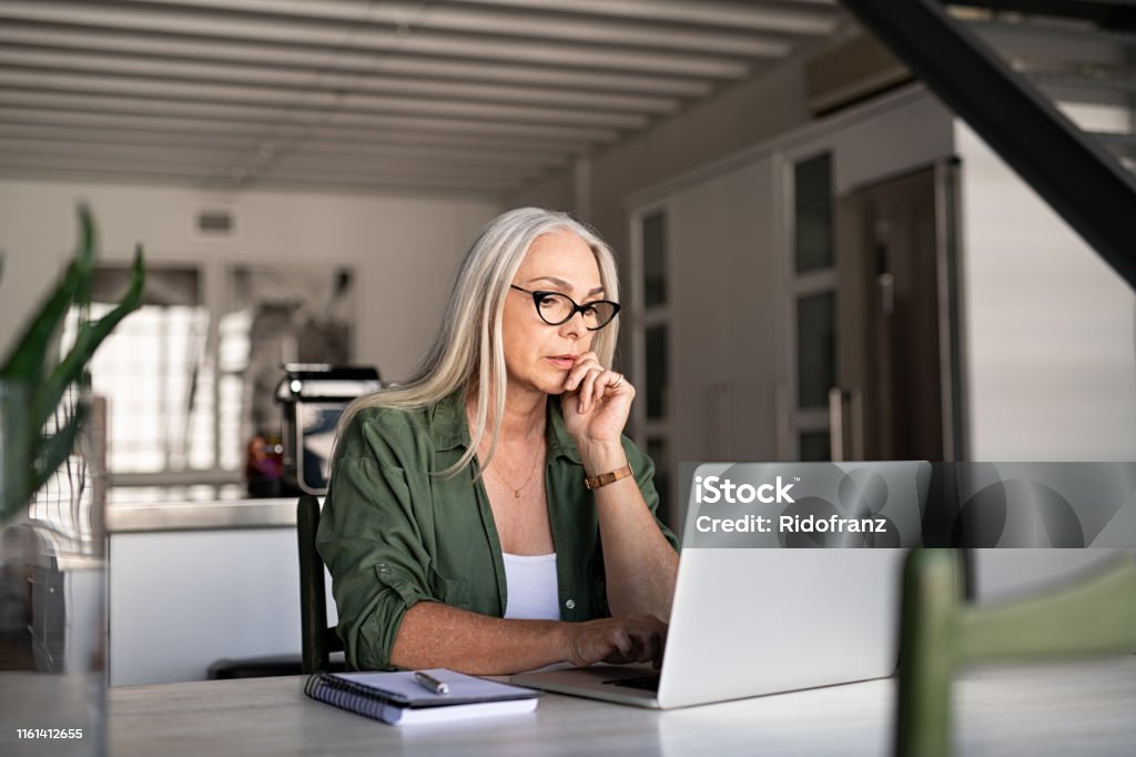 Worried senior woman using laptop Focused old woman with white hair at home using laptop. Senior stylish entrepreneur with notebook and pen wearing eyeglasses working on computer at home. Serious woman analyzing and managing domestic bills and home finance. Laptop Stock Photo