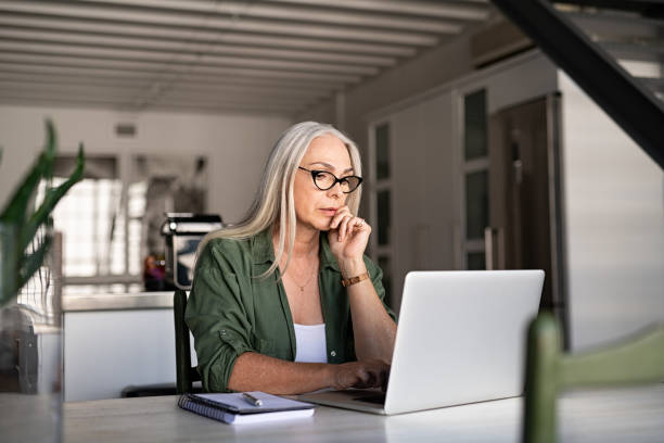 Worried senior woman using laptop Focused old woman with white hair at home using laptop. Senior stylish entrepreneur with notebook and pen wearing eyeglasses working on computer at home. Serious woman analyzing and managing domestic bills and home finance. grave stock pictures, royalty-free photos & images