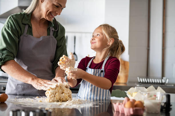 Smiling grandmother and happy child kneading dough Playful grandmother and smiling child kneading cookie dough in kitchen. Happy granddaughter in apron with messy dough hands looking at senior woman while preparing a pie at home. Smiling little girl helping grandma making biscuits on kitchen counter. baking bread photos stock pictures, royalty-free photos & images