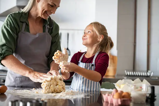 Playful grandmother and smiling child kneading cookie dough in kitchen. Happy granddaughter in apron with messy dough hands looking at senior woman while preparing a pie at home. Smiling little girl helping grandma making biscuits on kitchen counter.