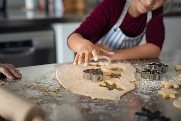 Closeup of child hands cutting cookies from dough. Little girl in apron pressing cookie cutter for shaping dough. Girl baking heart shaped christmas biscuits in the kitchen.