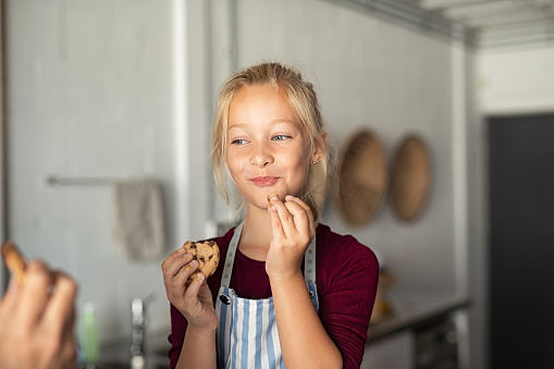 Cheerful little girl tasting chocolate chip cookies. Girl wearing apron and eating chocolate chip cookie in kitchen with grandmother. Happy girl enjoying self made biscuits at home with funny expression.