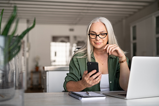 Happy senior woman using mobile phone while working at home with laptop. Smiling cool old woman with white hair wearing eyeglasses sitting on chair at table and messaging with smartphone. Beautiful stylish elderly lady browsing site on cellphone.