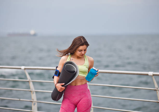 la jeune femme d'ajustement dans le sportwear retient le tapis de yoga et la bouteille d'eau dans sa main et posant à l'extérieur sur la plage - aerobics beautiful bottle body photos et images de collection