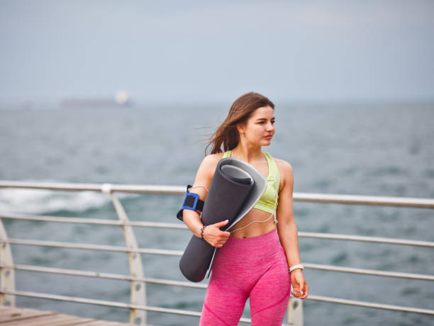 jeune femme athlétique dans le sportwear retient le tapis de yoga dans sa main et posant à l'extérieur sur la plage - aerobics beautiful bottle body photos et images de collection