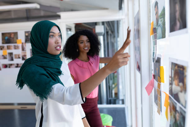 Female graphic designers discussing over photographs in office Side view of young diverse female graphic designers discussing over photographs in office. This is a casual creative start-up business office for a diverse team sales executive stock pictures, royalty-free photos & images