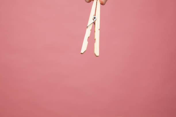 Hand holding a wooden clamp to spread clothes. The clamp is horizontal and vertical, open and closed, on a plain pink background.