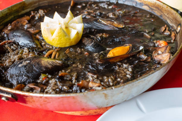 primo piatto di riso nero con calamari "u2013in ink"u2013 cozze, vongole e altri frutti di mare. presentato a paellera. - seafood lobster paella prepared shellfish foto e immagini stock