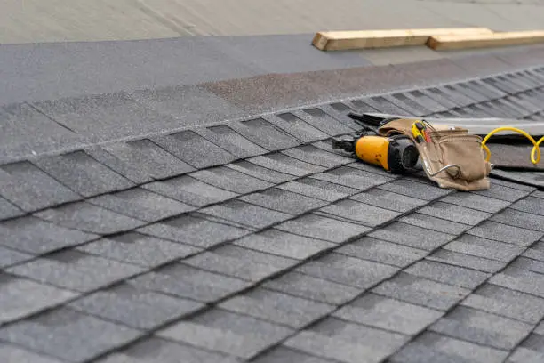 Photo of Photo of toolbelt with instrument and nail gun lying on asphalt or bitumen shingle on top of the new roof under construction residential house or building