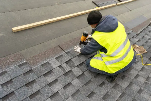 Photo of Qualified workman in uniform work wear using air or pneumatic nail gun and installing asphalt or bitumen shingle on top of the new roof under construction residential building