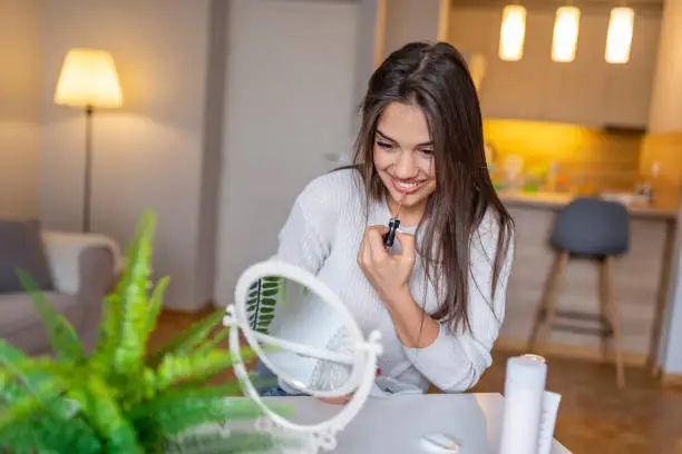 Portrait of a beautiful woman, dyes her lips lipstick, looking in the mirror. Beautiful girl is using a lip-gloss while looking into the mirror at home. Woman lipstick mirror