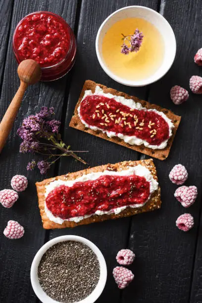 Healthy sugar free dessert and herbal tea. Raspberry chia jam on crispy bread and cream cheese with frozen berries on black background top view