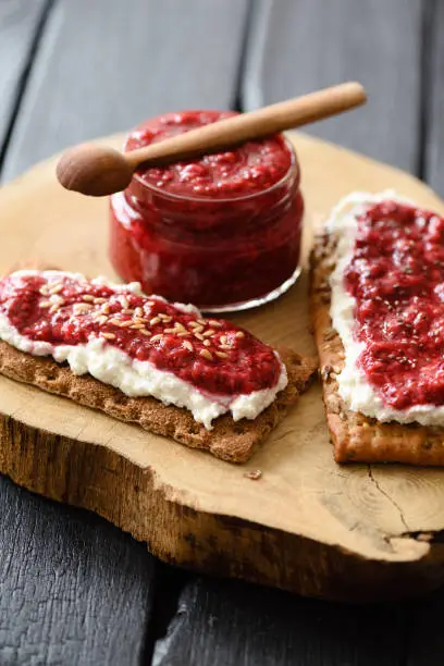 Healthy sugar free preserves. Raspberry chia jam with flax seeds on crispy breads and cream cheese on black background high angle view