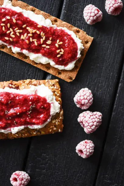 Healthy sugar free dessert. Raspberry chia jam and flax seeds on crispy bread and cream cheese with frozen berries on black background top view