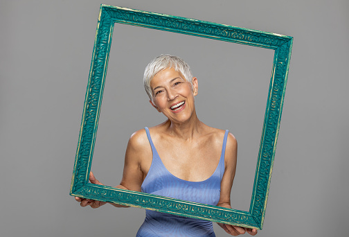 Portrait of a beautiful elderly woman, smiling, holding picture frame, isolated on a grey background.