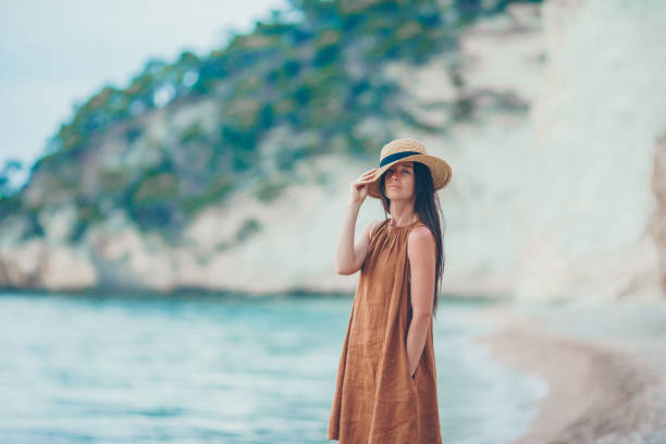 Young beautiful woman during tropical beach vacation. Girl in hat and brown dress relax on the beach in sunset