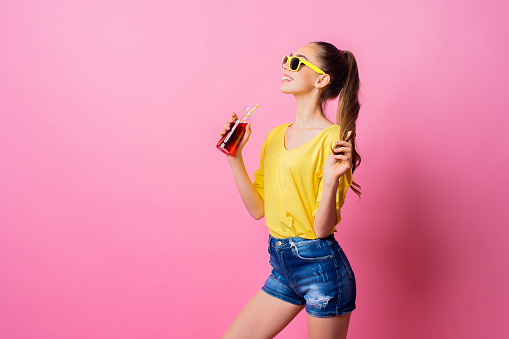 Teenage female in sunglasses standing in studio holding bottle and drinking red beverage through straw on pink background