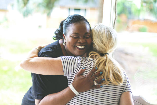 adult african american female e caucasica davanti alla porta di residenza salutarsi con un abbraccio - vicino di casa foto e immagini stock