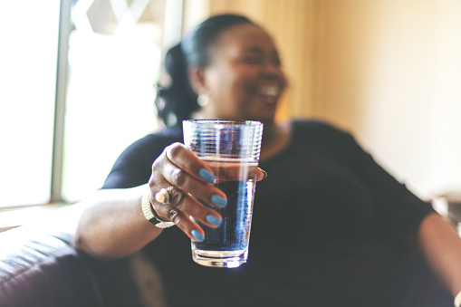 istock Relaxed Smiling Adult African American Female on living room sofa offering glass of water to unpictured guest with window and lamp back-lighting 1161471096