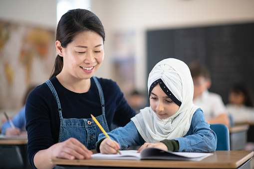A female teacher of Asian descent helps her female  Muslim student write in her book. She is kneeling beside her while her student sits at her desk focused.